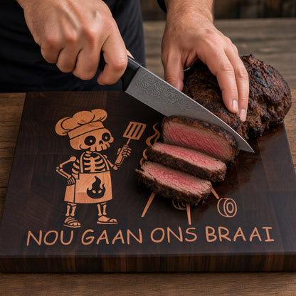 A premium everyday end-grain cutting board with Brass feet.

Materials: Base is walnut wood with a hard maple inlay.

Design: With a carved skeleton chef grilling next to a charcoal grill, with the text 'NOU GAAN ONS BRAAI' below.

Uses:
- Chopping Board
- Cheese or Meat Board
- Charcuterie Board
- Biltong Board

Note: Wood is a natural material, and the received product will have wood color and grain variations compared to this picture.